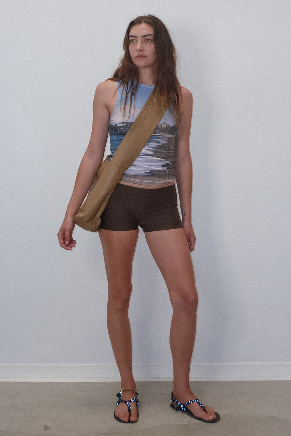 woman wearing low rise brown swim shorts, blue printed mt fuji organic cotton tank top and light brown minimalist leather cross body bag.