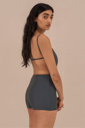 Model wearing blueish grey Terrain One Piece shorty surf suit — sustainable swimwear designed for support and comfort in the surf, flattering one piece surf suit for active women.