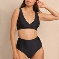 A high-waist black sustainable swimwear bikini bottom with minimalist design and moderate coverage, modelled by a woman in a matching bikini top.