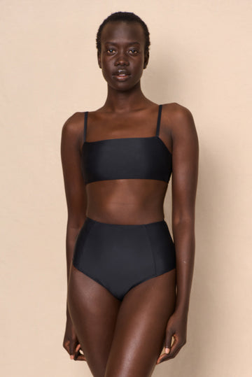 A high-waist black sustainable swimwear bikini bottom with minimalist design and moderate coverage, modelled by a woman in a matching bikini top.